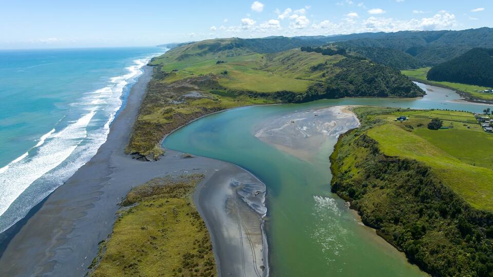 A coastal landscape with a river curving through green hills, a beach, and a clear blue ocean.