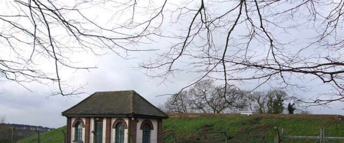 A small brick building with a pitched roof and arched windows, surrounded by a grassy field and a fence.