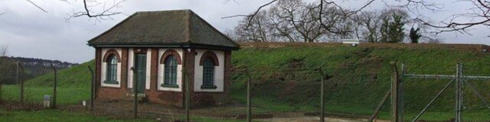 A small brick building with a pitched roof and arched windows, surrounded by a grassy field and a fence.