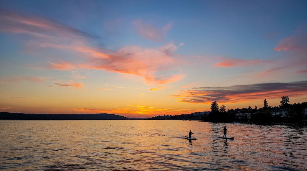 A sunset over a calm lake with paddle boats, a distant shoreline with trees, and a clear sky with clouds.
