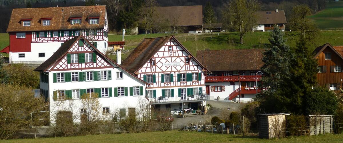 A traditional half-timbered house with green shutters, surrounded by a rural landscape with fields and distant hills.