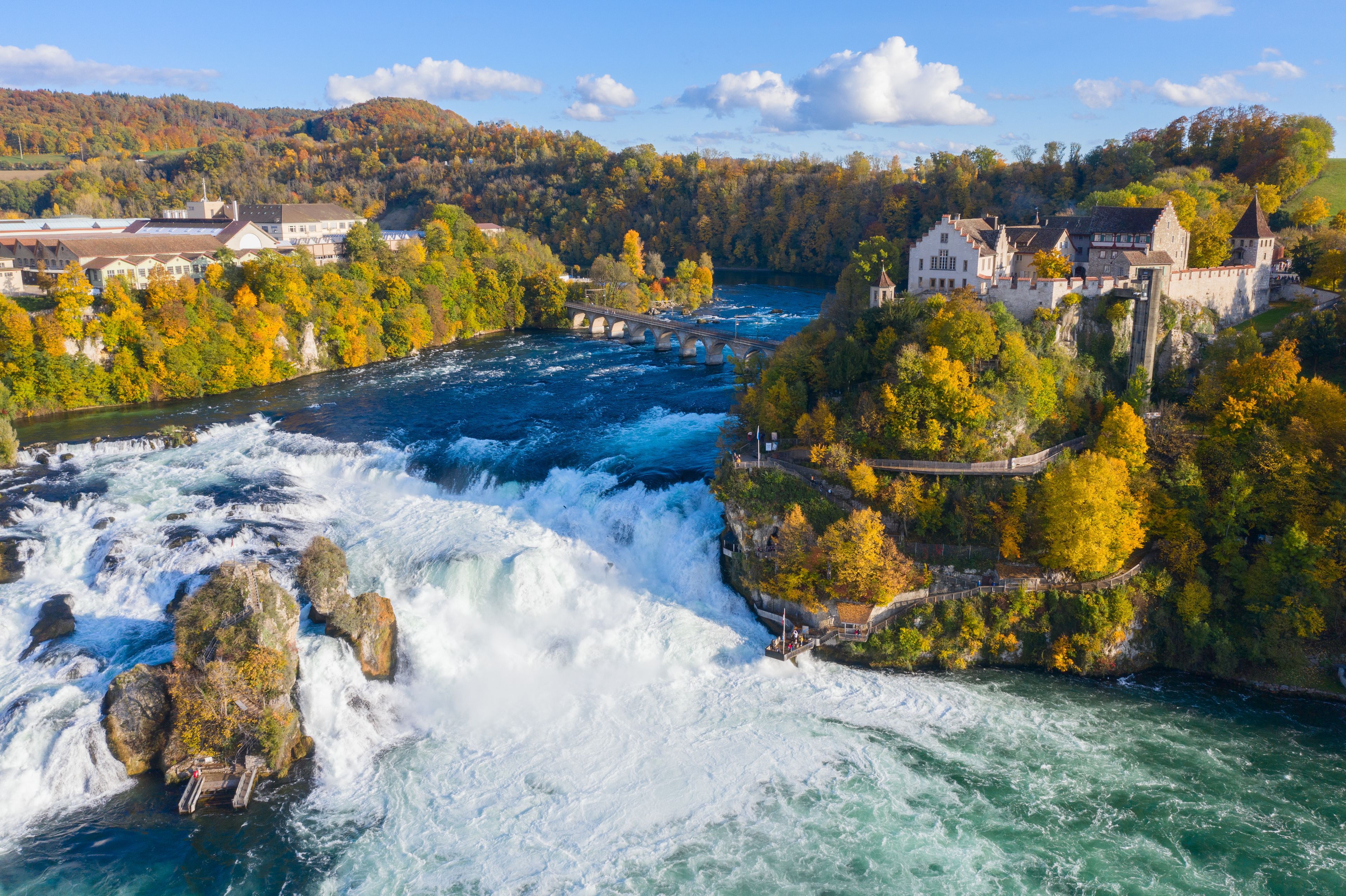A river with a waterfall, surrounded by autumn-colored trees and historic buildings.