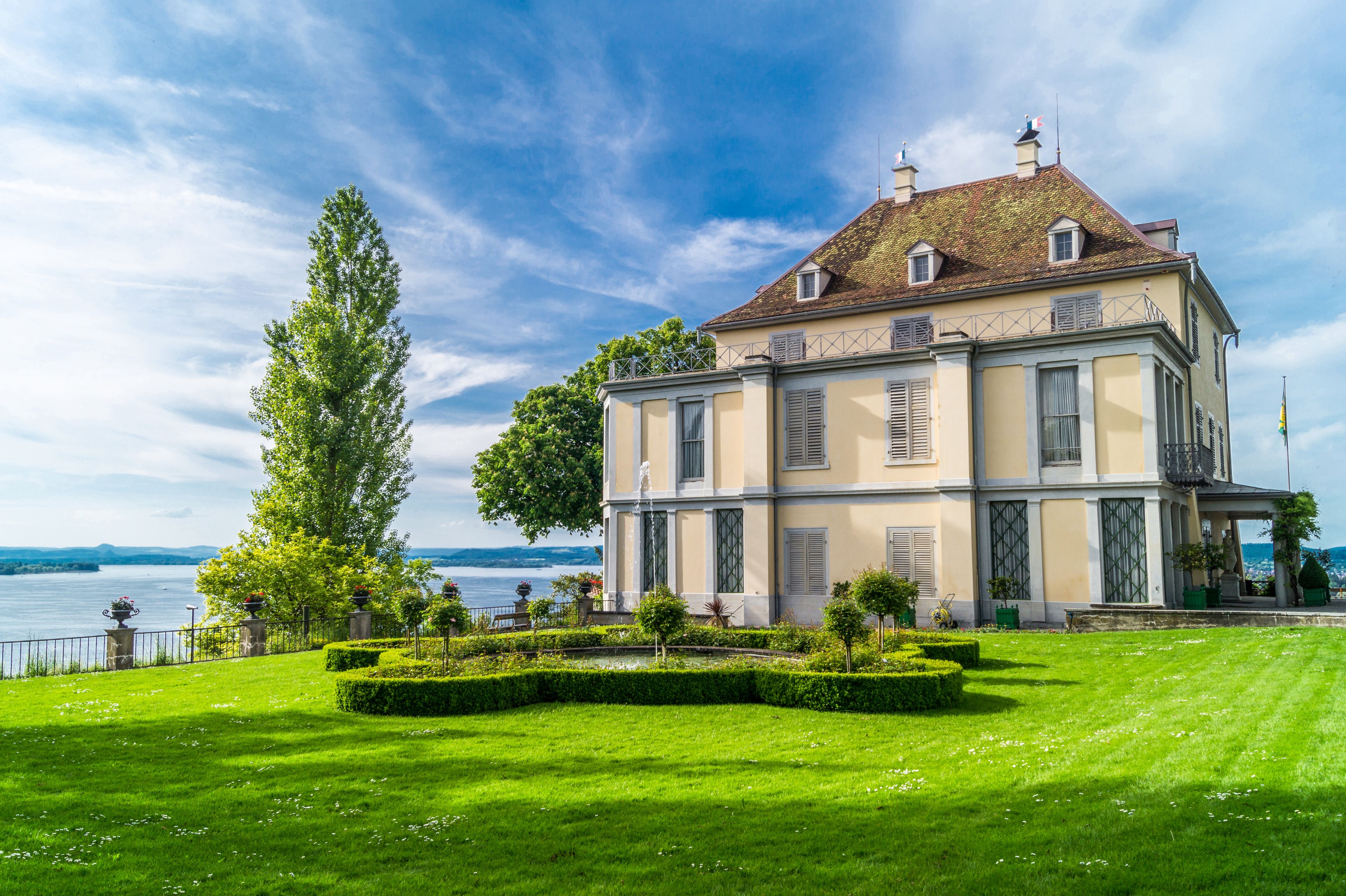 A large, historic building with a symmetrical facade, a well-manicured lawn, and a body of water in the background.