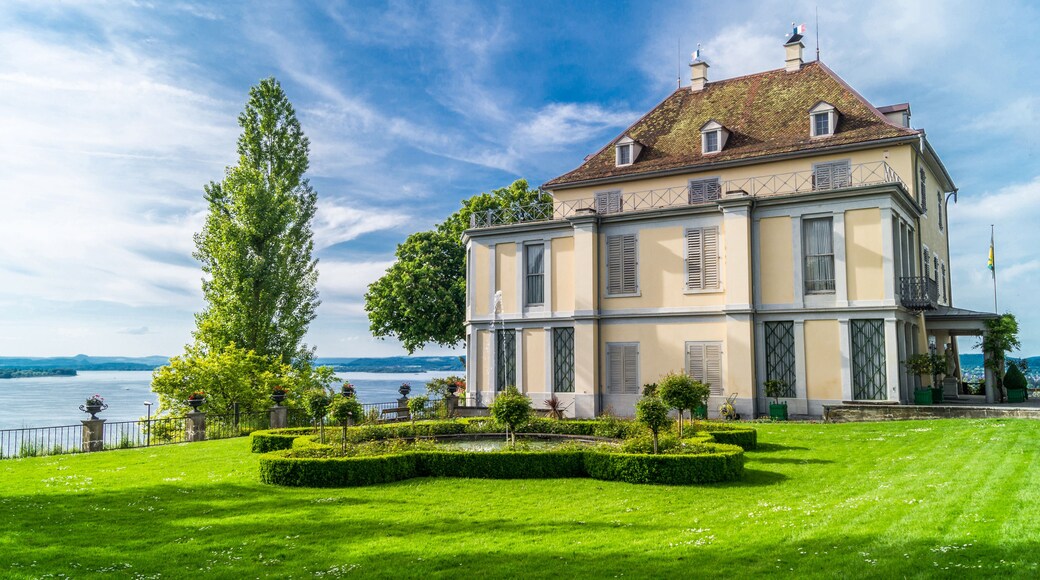 A large, historic building with a symmetrical facade, a well-manicured lawn, and a body of water in the background.
