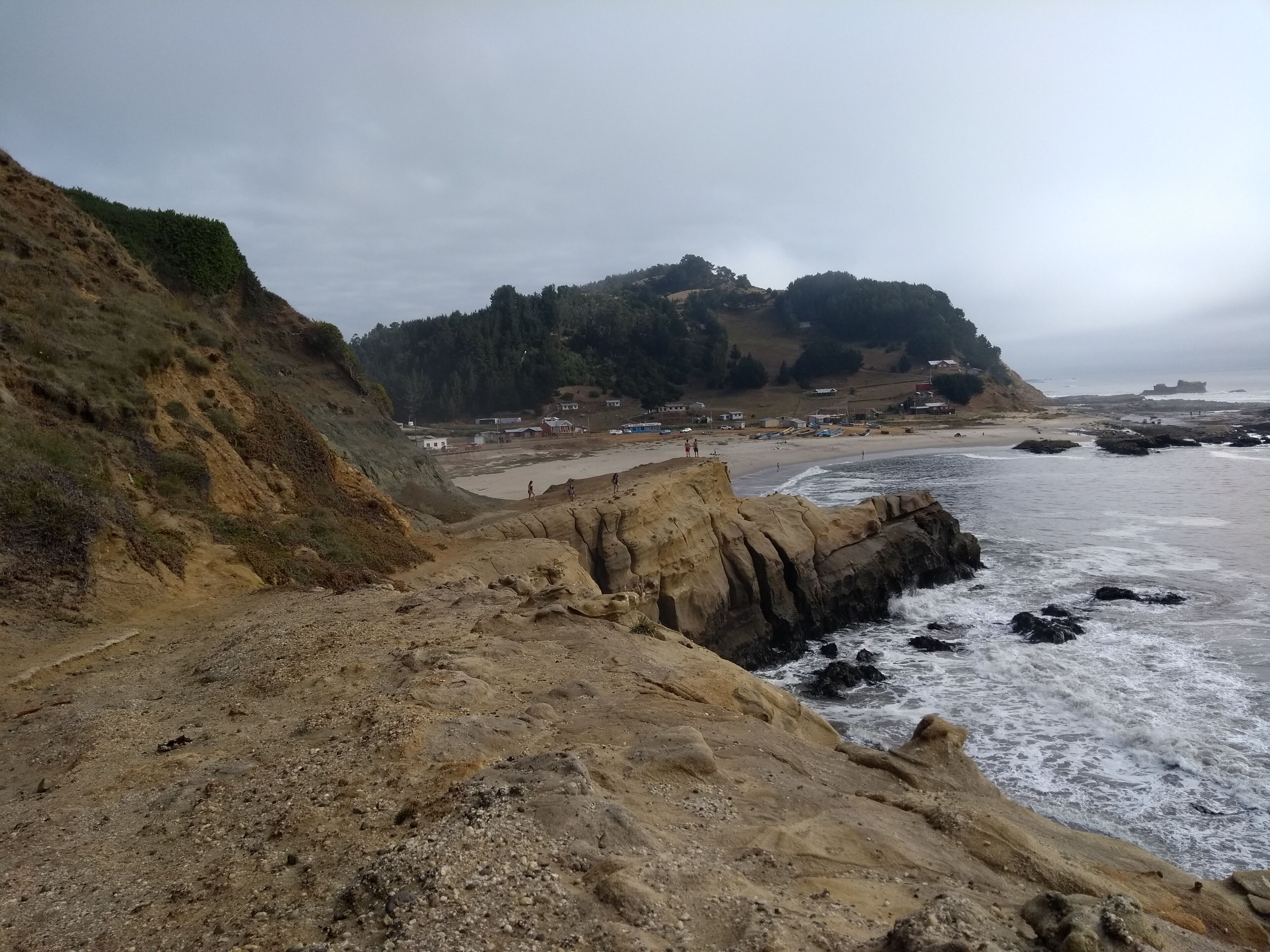A coastal landscape with rocky cliffs, a sandy beach, and a forested hill in the background.