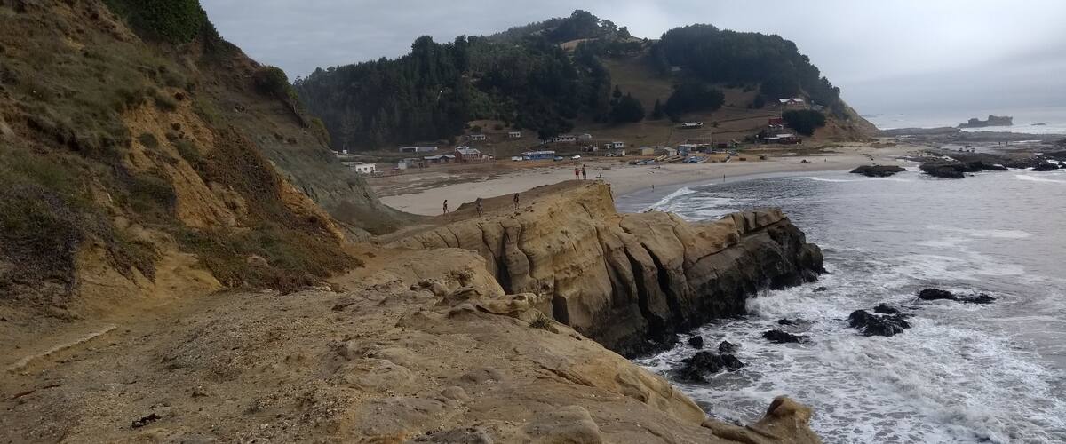 A coastal landscape with rocky cliffs, a sandy beach, and a forested hill in the background.