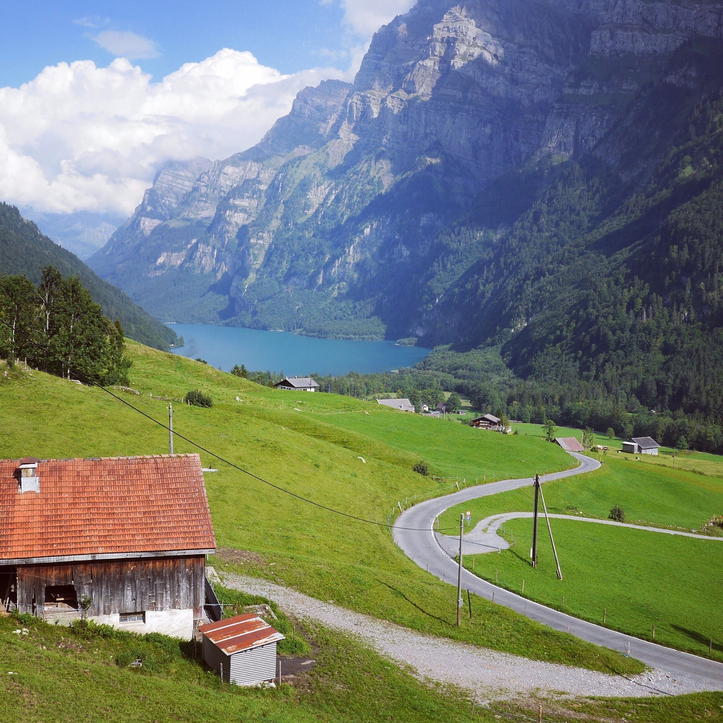 A rural landscape with a winding road, a barn, and a lake surrounded by mountains.