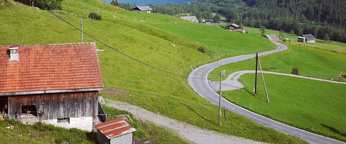 A rural landscape with a winding road, a barn, and a lake surrounded by mountains.