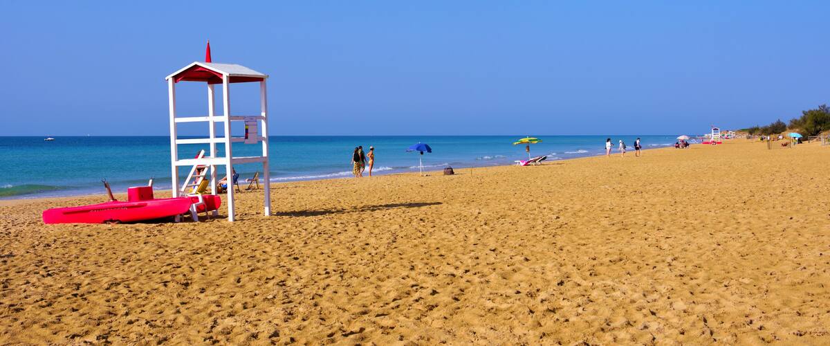 A sandy beach with a lifeguard tower, red lifeguard stand, and a few people in the distance.