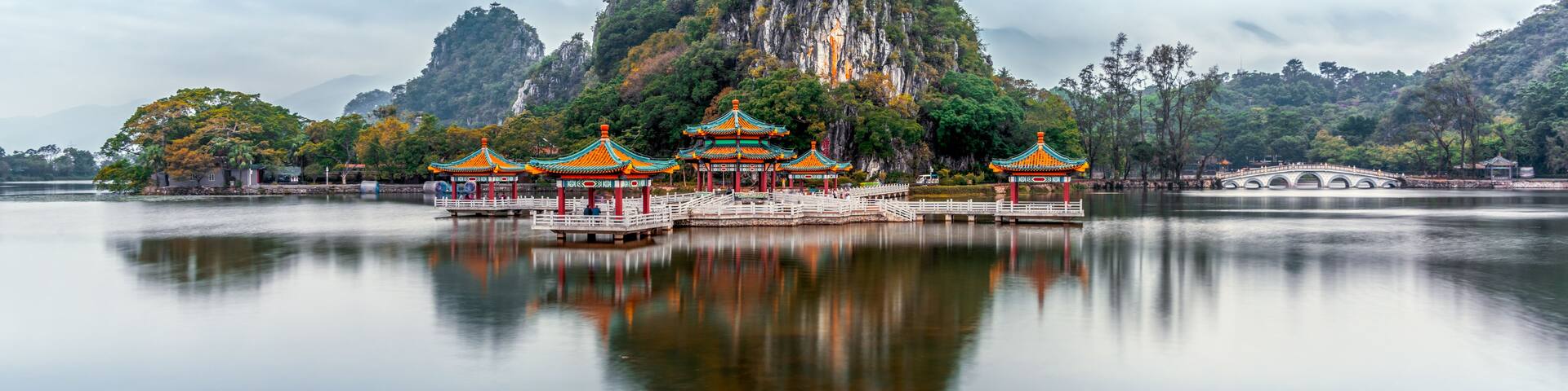 A traditional Chinese temple complex with multiple pagodas, a large mountain, and a calm lake.