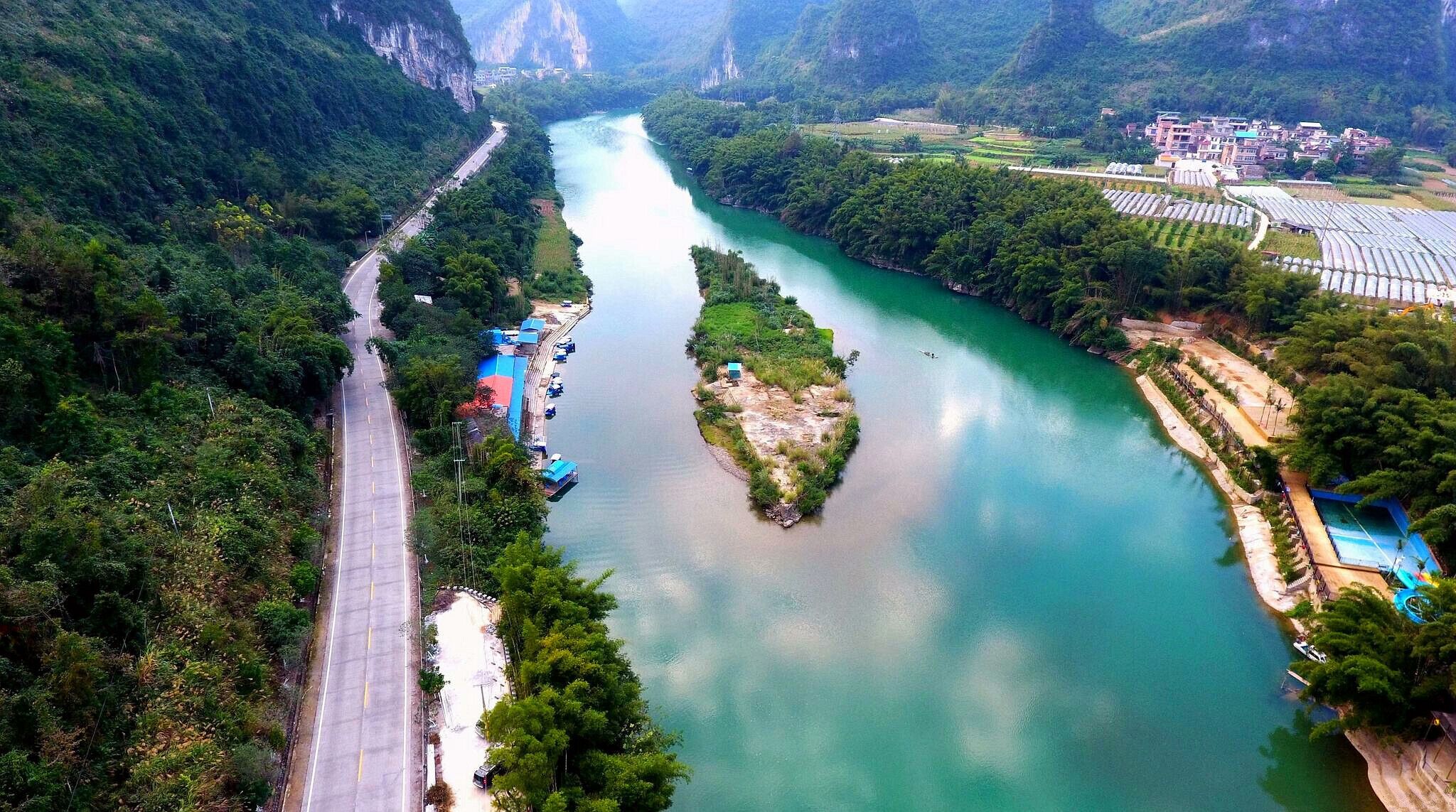 A river winding through a mountainous landscape with a road running alongside it. There are buildings and a small island in the river.
