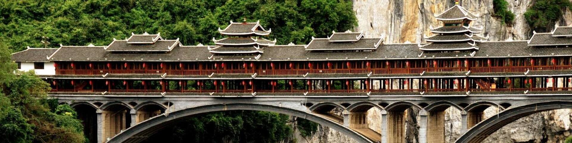 A traditional Chinese bridge with multiple arches spanning a river, surrounded by lush greenery and rocky cliffs.