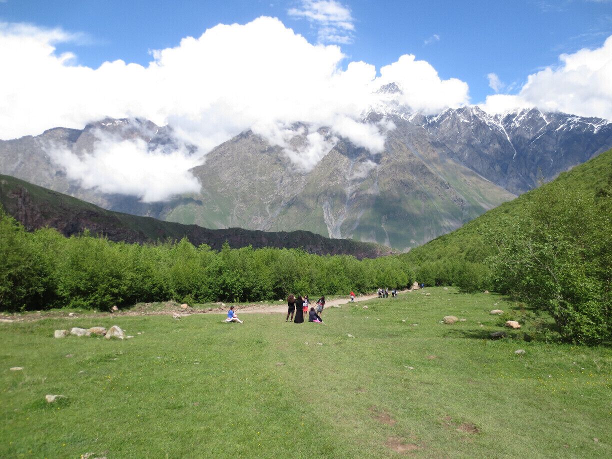 Un valle cubierto de hierba con gente caminando y sentada, rodeado de montañas con cumbres nevadas y nubes.