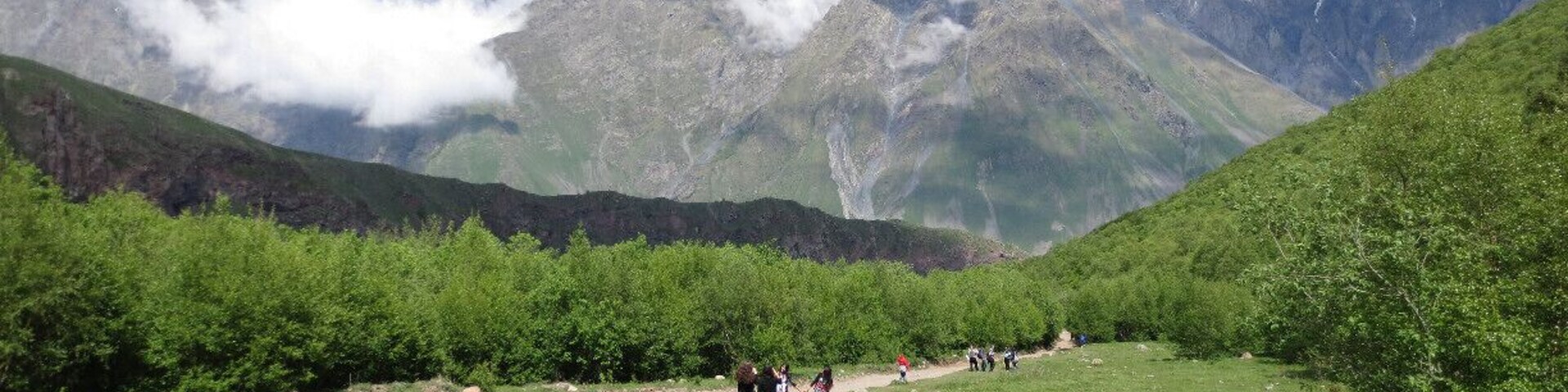 Un valle cubierto de hierba con gente caminando y sentada, rodeado de montañas con cumbres nevadas y nubes.