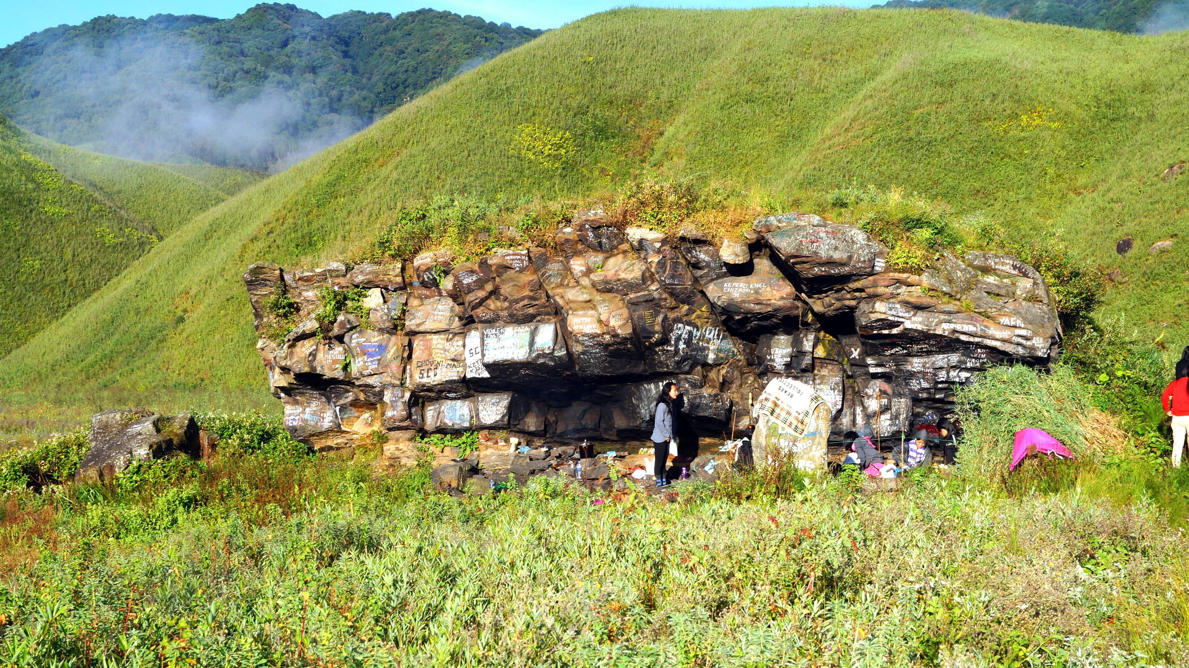 A large rock formation with inscriptions and graffiti, surrounded by lush green hills and vegetation.