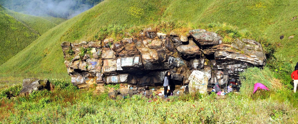 A large rock formation with inscriptions and graffiti, surrounded by lush green hills and vegetation.