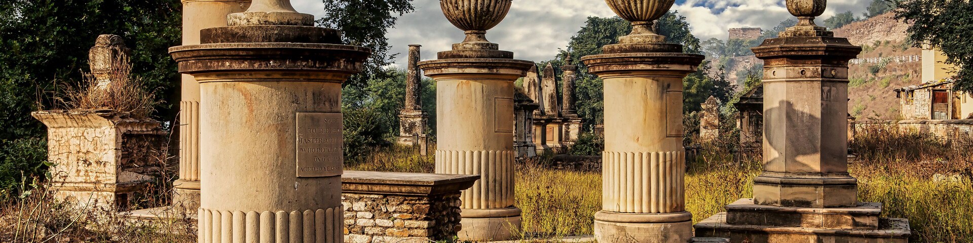 Columnas y urnas de piedra antiguas en un jardín descuidado con vegetación exuberante.