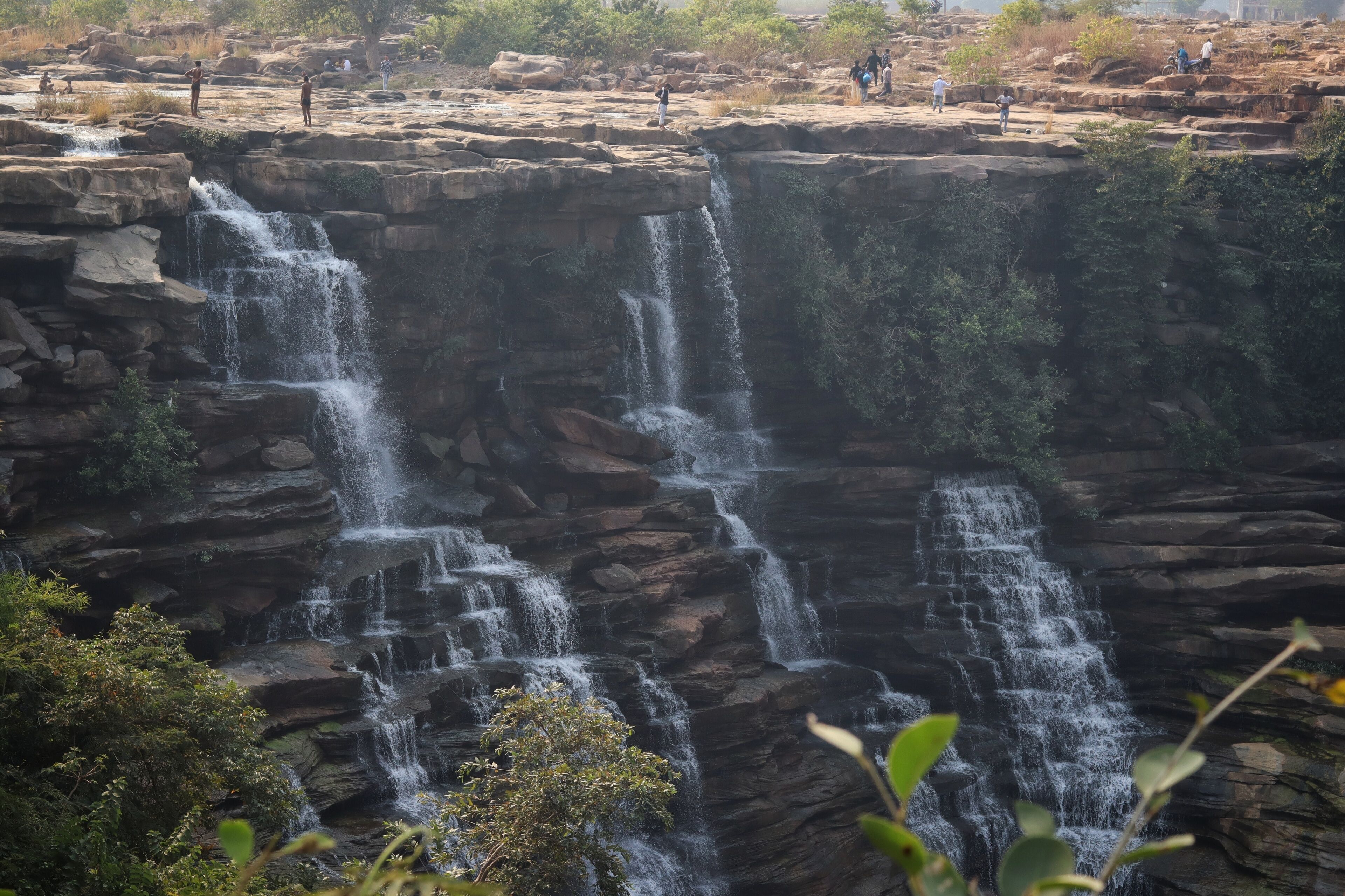 A multi-tiered waterfall cascading over rocky terrain with people observing from the top.