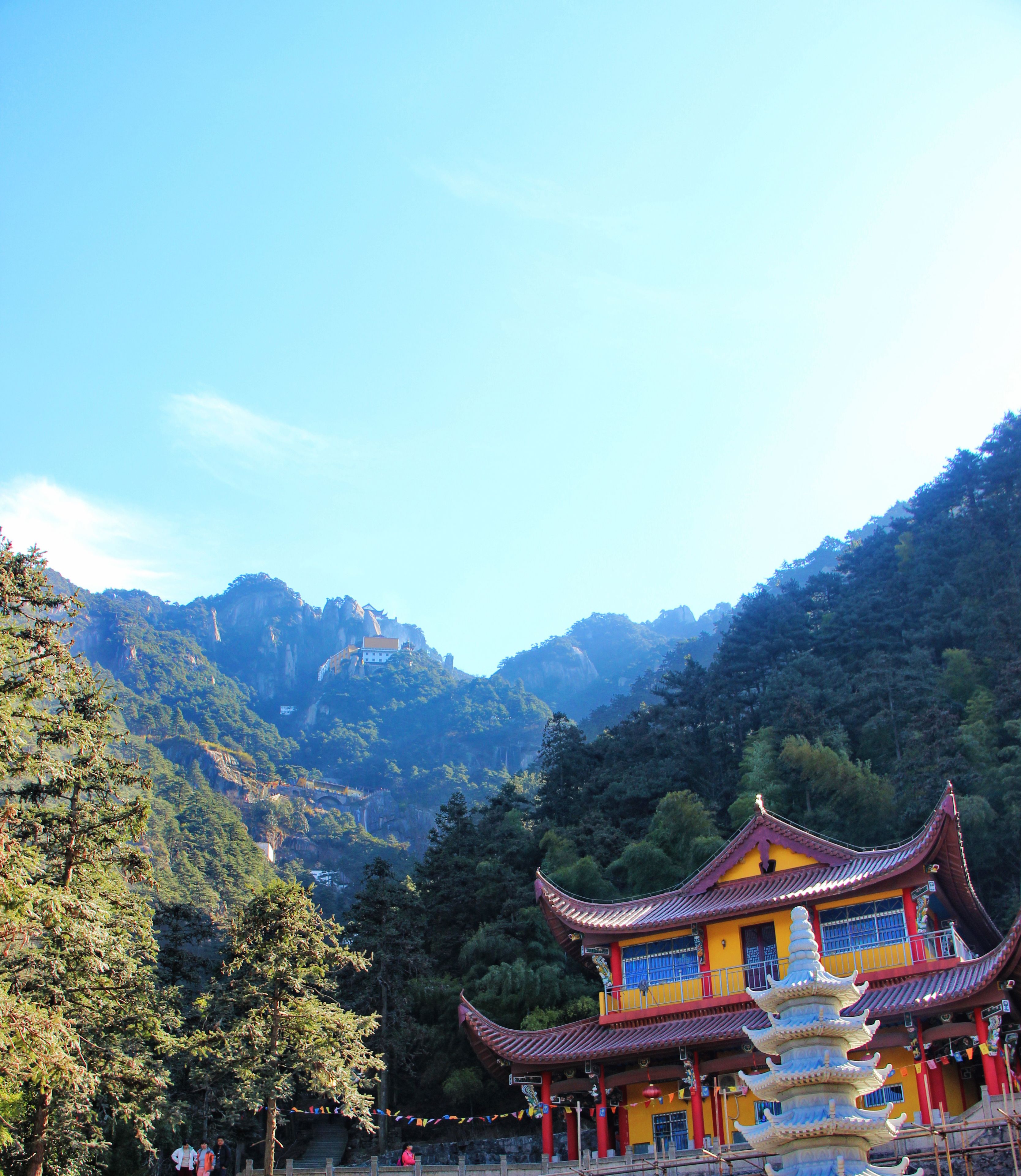 Un templo chino tradicional con una pagoda de varios niveles, rodeado de montañas verdes exuberantes y un cielo azul despejado.
