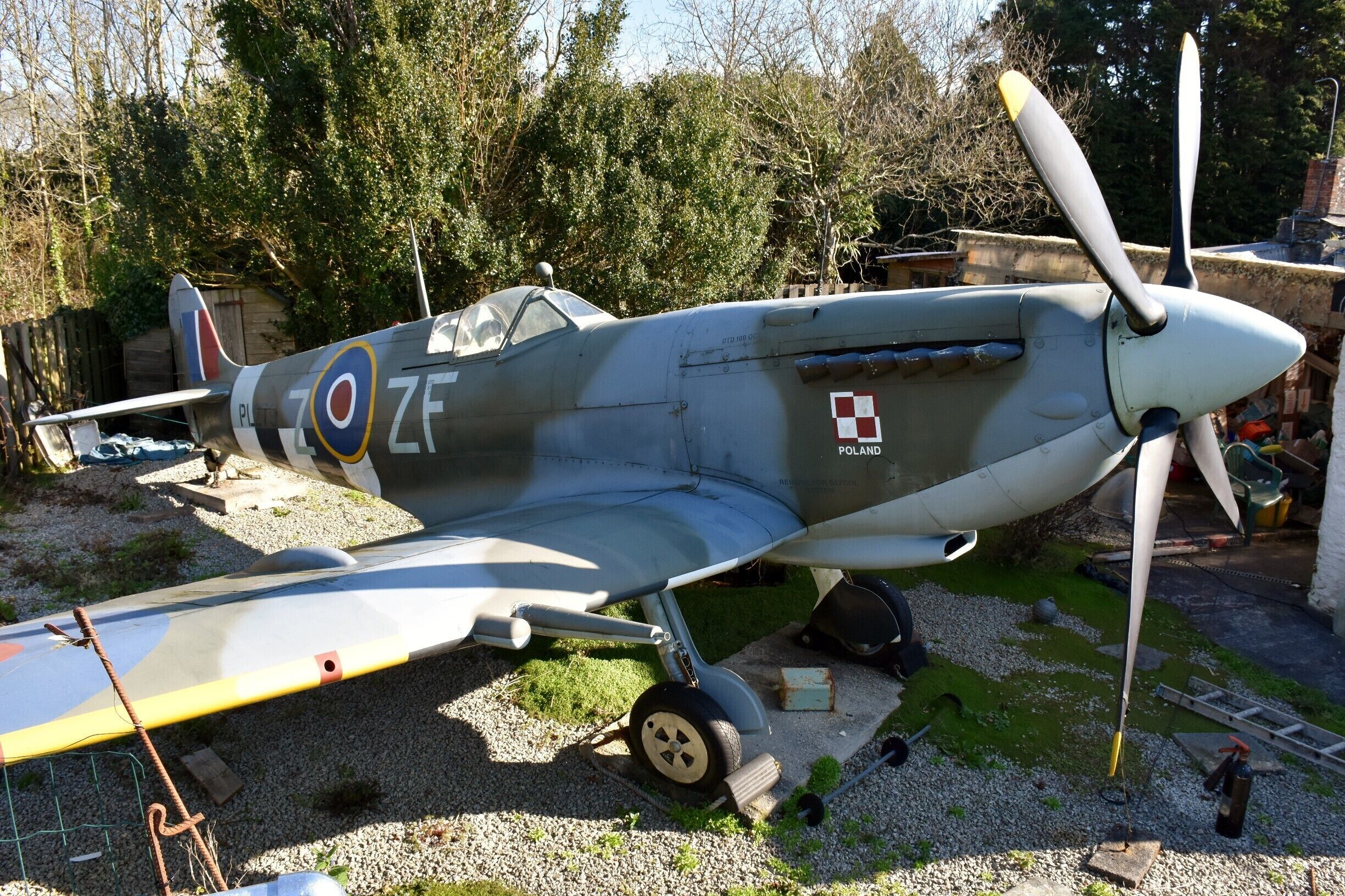A vintage military aircraft with a propeller, parked outdoors with trees and a building in the background.