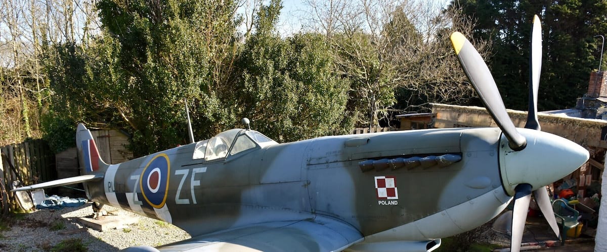 A vintage military aircraft with a propeller, parked outdoors with trees and a building in the background.