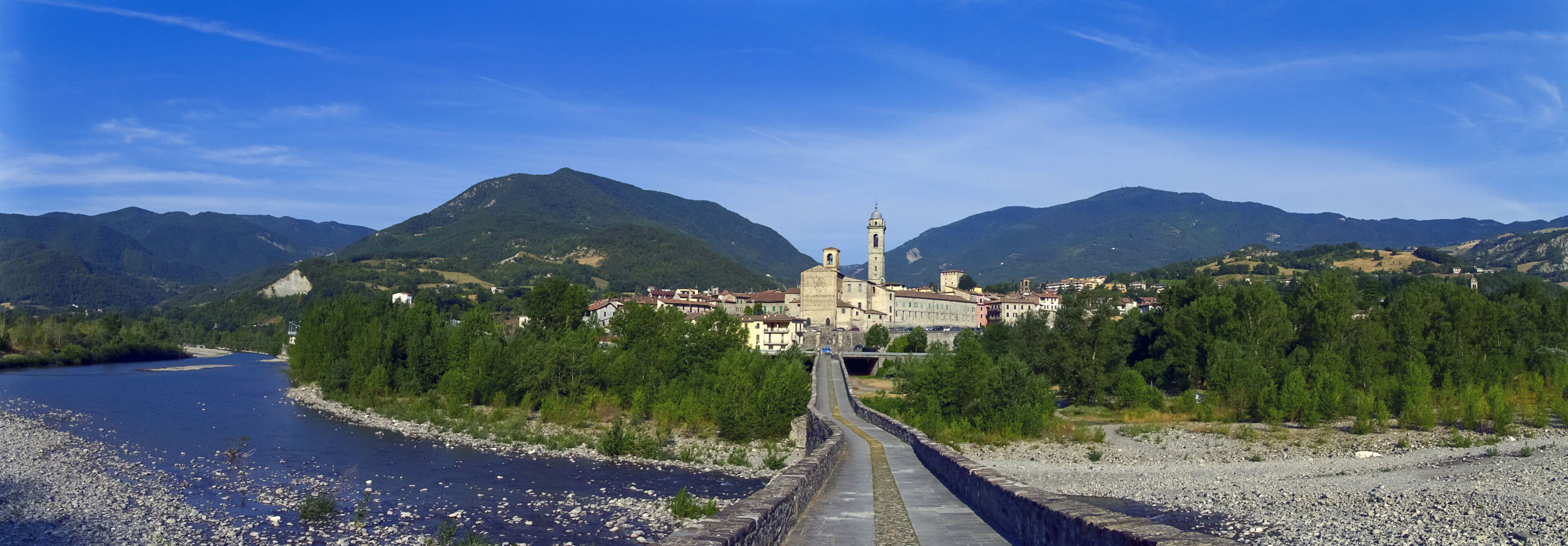 A river with a stone bridge, a town with historic buildings, and mountains in the background.