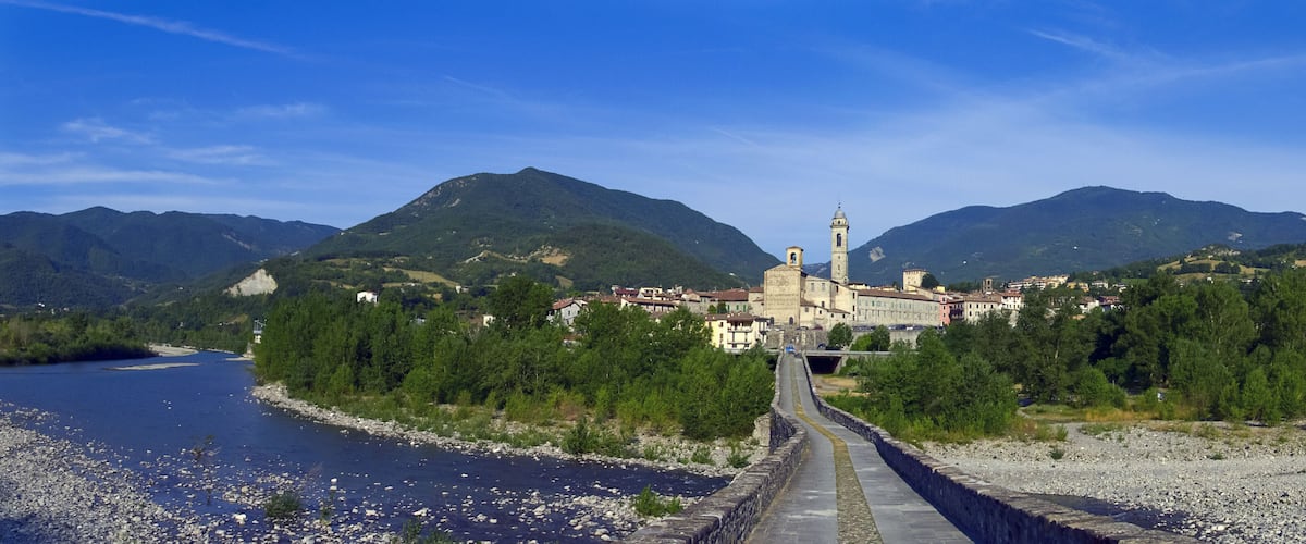 A river with a stone bridge, a town with historic buildings, and mountains in the background.