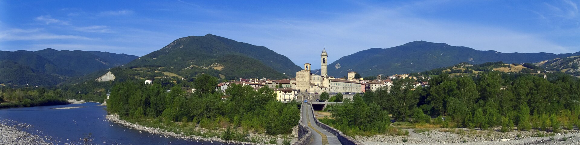A river with a stone bridge, a town with historic buildings, and mountains in the background.