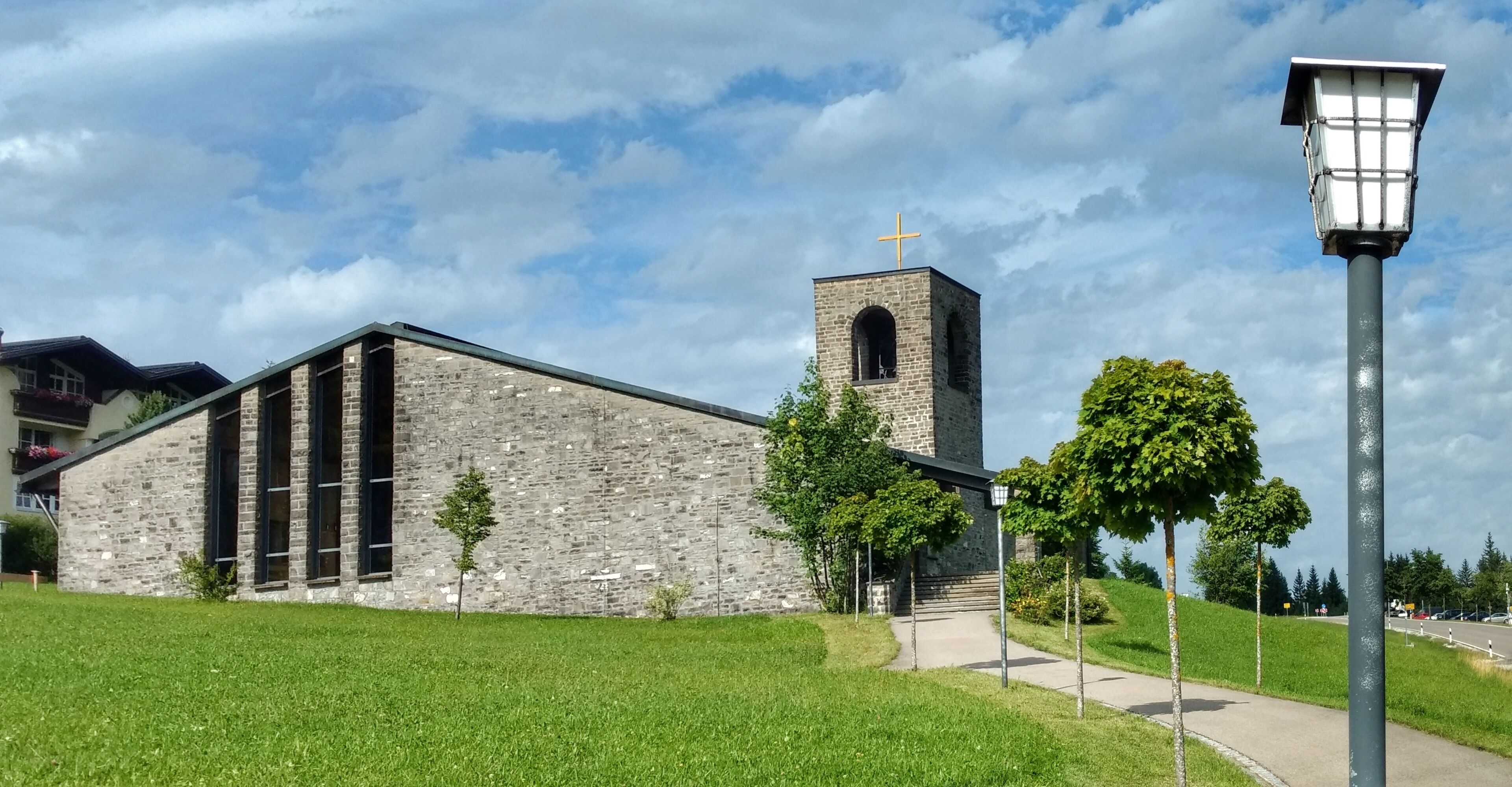A stone church with a bell tower and a cross on top, surrounded by green grass and trees.