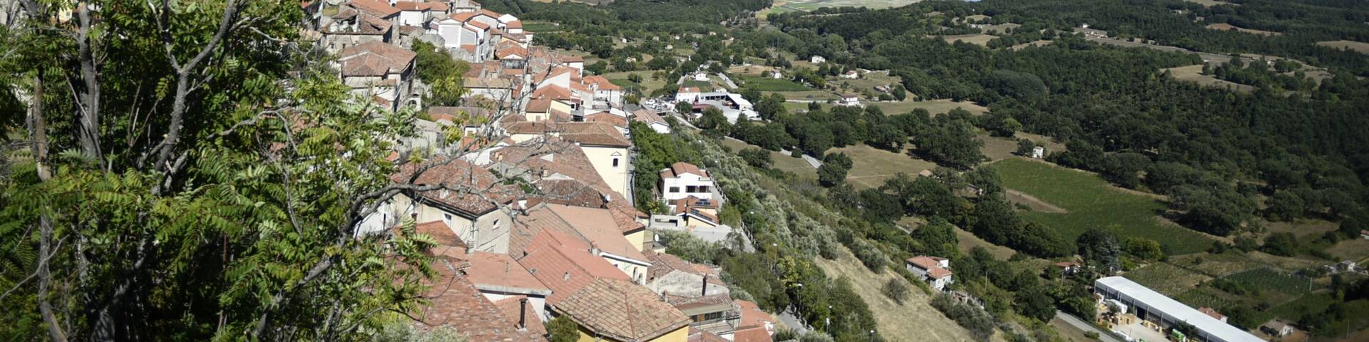 A coastal town with terracotta-roofed buildings, a church, and a view of the sea and hills in the distance.