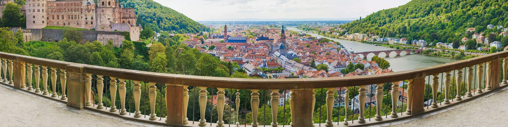 A historic castle overlooking a river, surrounded by lush greenery and a town with red-roofed buildings.