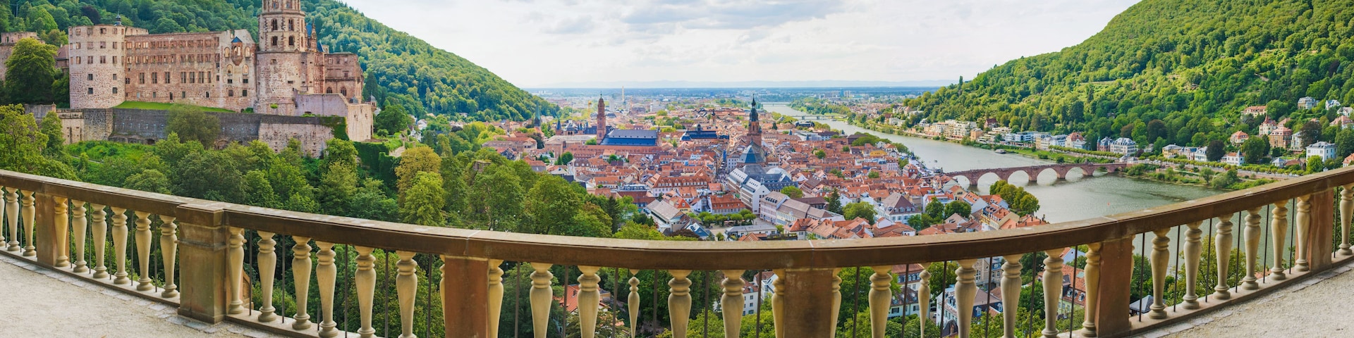 Eine historische Burg mit Blick auf einen Fluss, umgeben von üppiger Vegetation und einer Stadt mit roten Dächern.