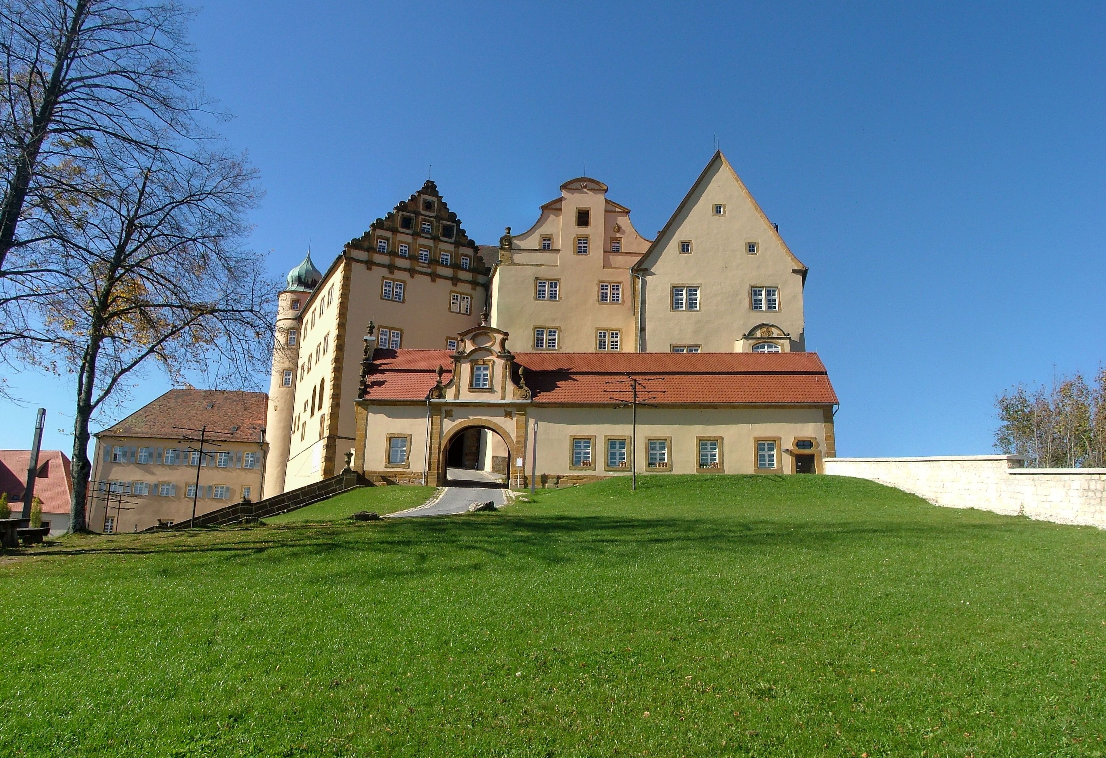 A large, historic building with a red roof and multiple towers, situated on a grassy hill.