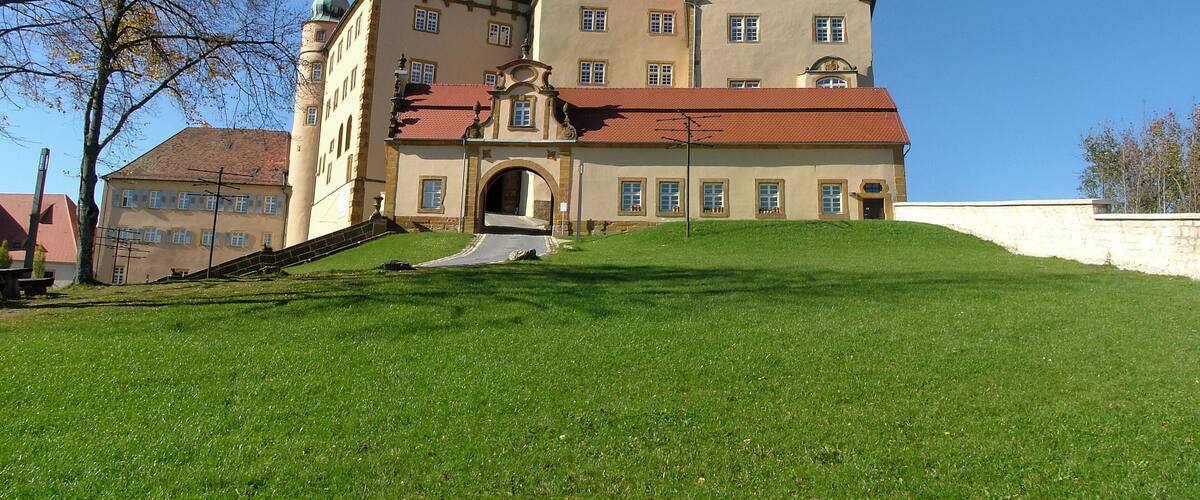 A large, historic building with a red roof and multiple towers, situated on a grassy hill.