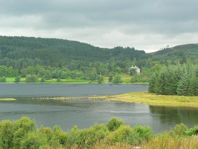 A lake surrounded by forested hills, a small house, and a grassy shoreline.