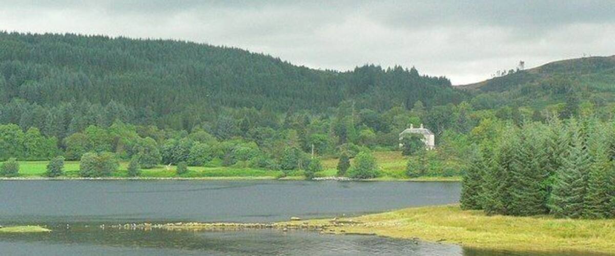 A lake surrounded by forested hills, a small house, and a grassy shoreline.