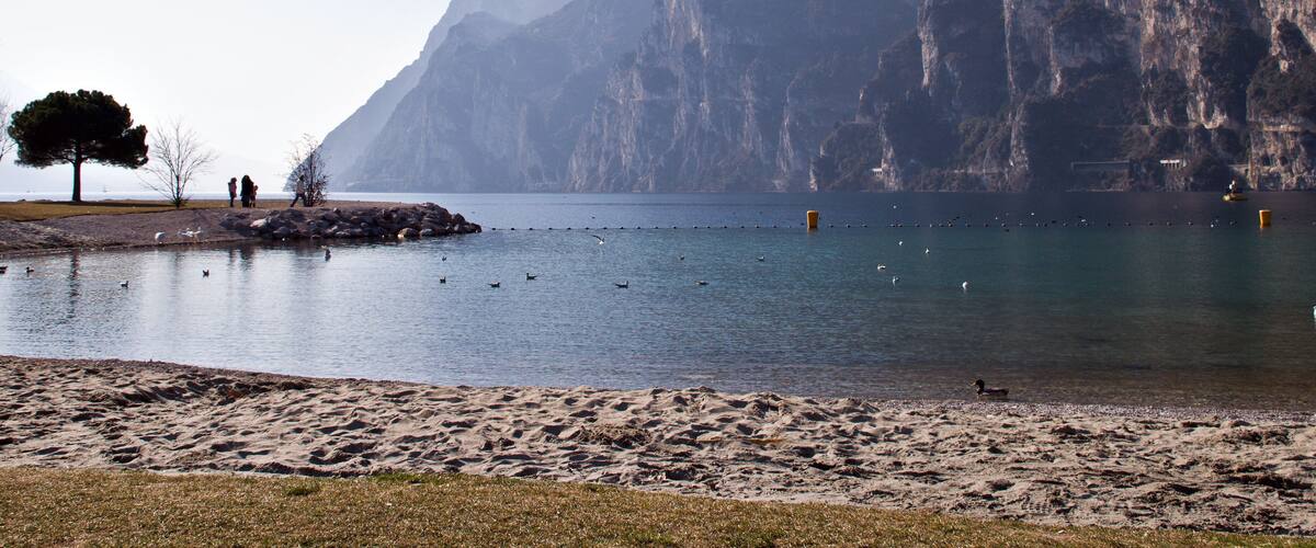 A calm lake with a sandy beach, a few people, and a large mountain in the background.