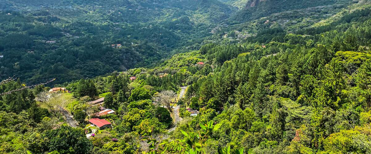 A lush valley with dense greenery, scattered houses, and distant mountains under a partly cloudy sky.