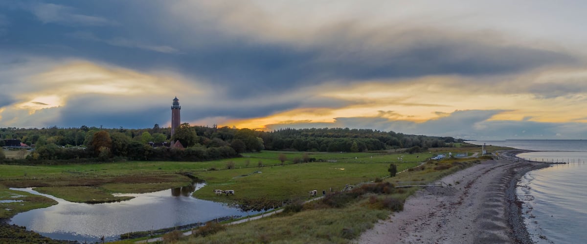 A coastal landscape with a lighthouse, grassy fields, a body of water, and a cloudy sky.