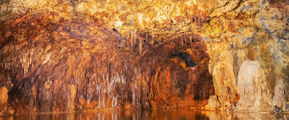 A cave with stalactites and stalagmites, a reflective pool, and a cave entrance.