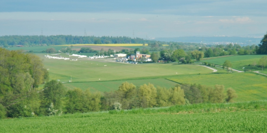 Paisatge rural amb camps verds, un petit grup d'edificis i una vila a la distància.