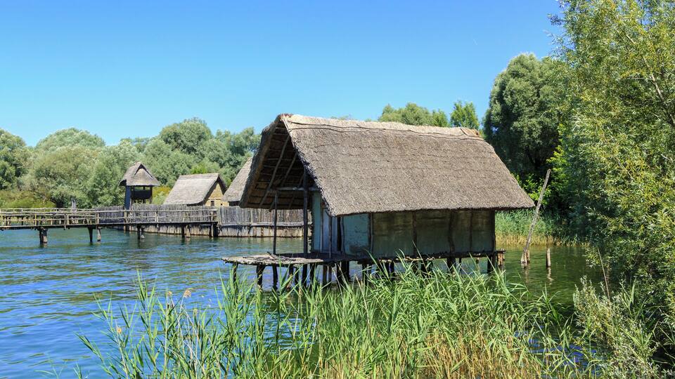 Thatched-roof huts on stilts over a calm river, surrounded by lush greenery.