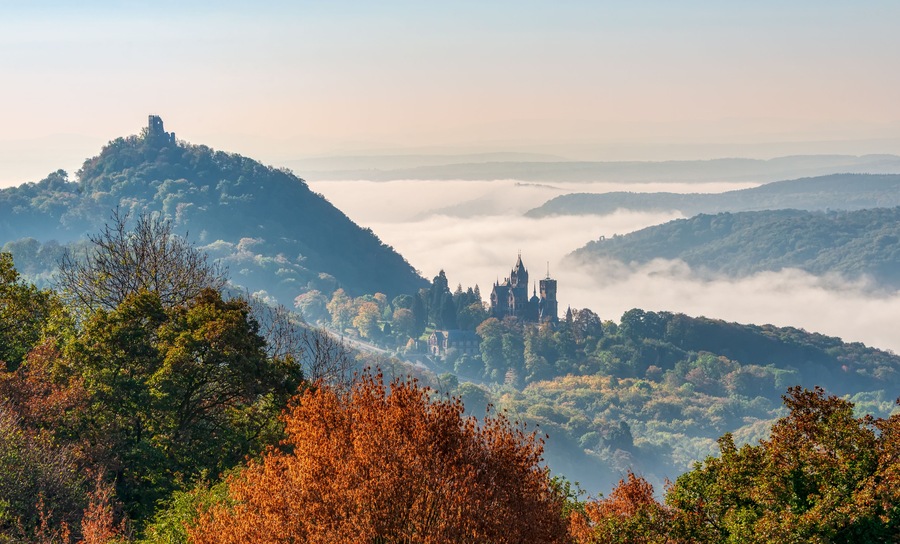 Un castillo en una colina rodeado de follaje otoñal y montañas cubiertas de niebla.