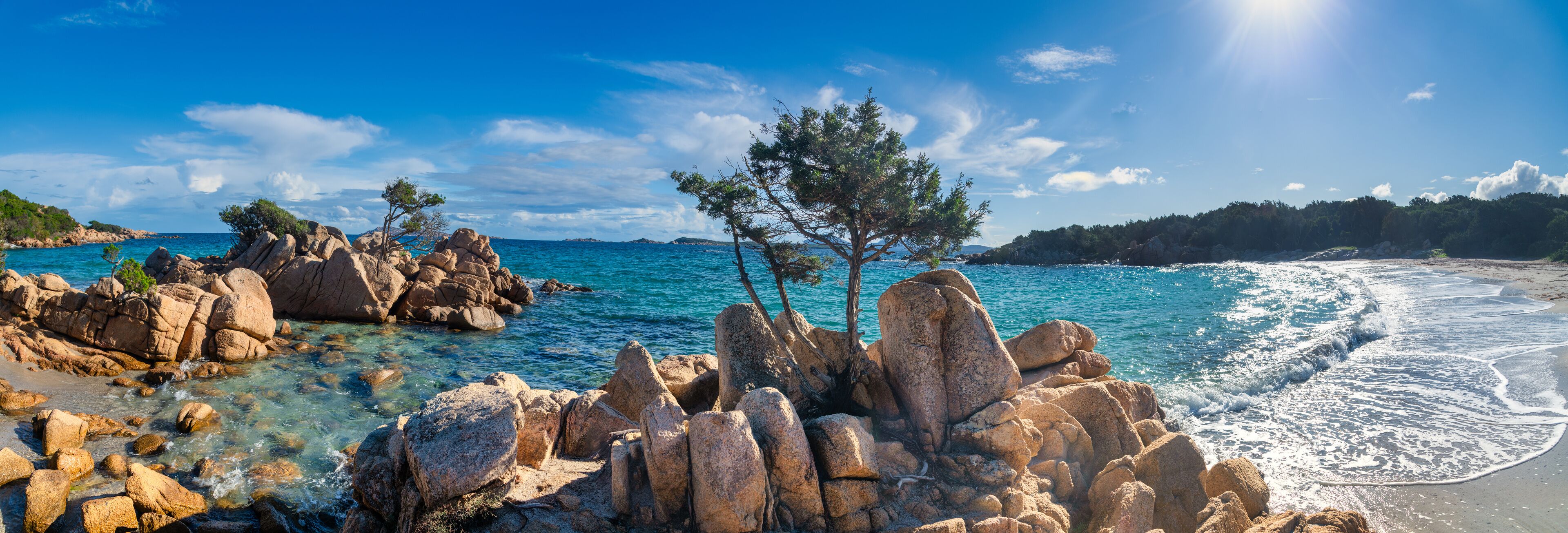 A rocky coastline with clear blue waters, a sandy beach, and a few trees.