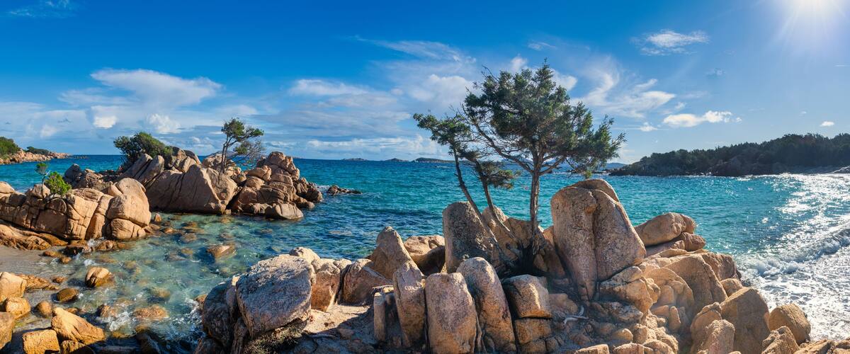 A rocky coastline with clear blue waters, a sandy beach, and a few trees.