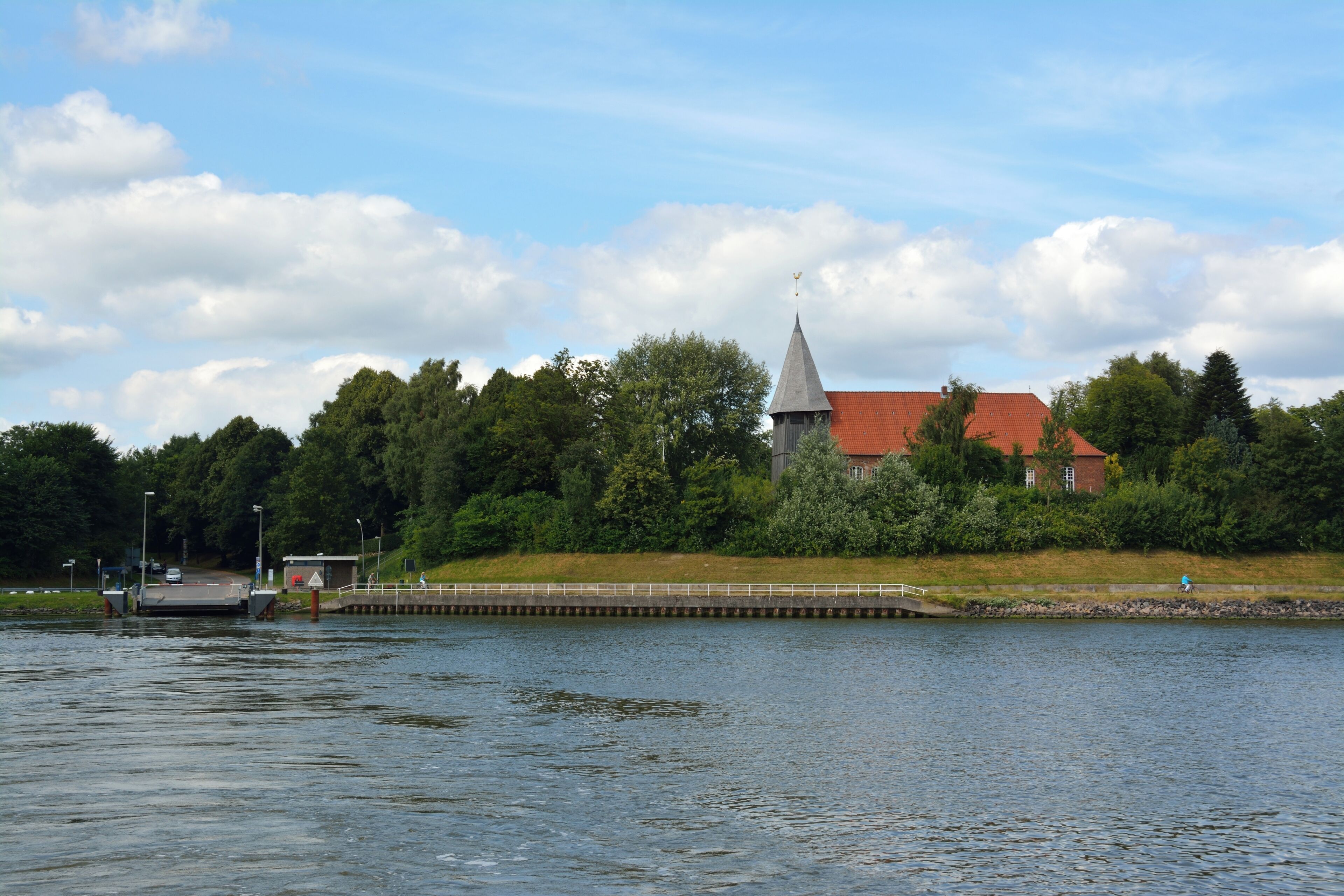A church with a tall steeple, a dock, and a bridge over a calm river.