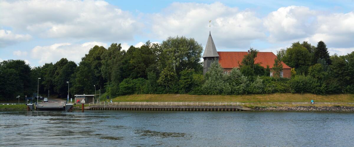 A church with a tall steeple, a dock, and a bridge over a calm river.