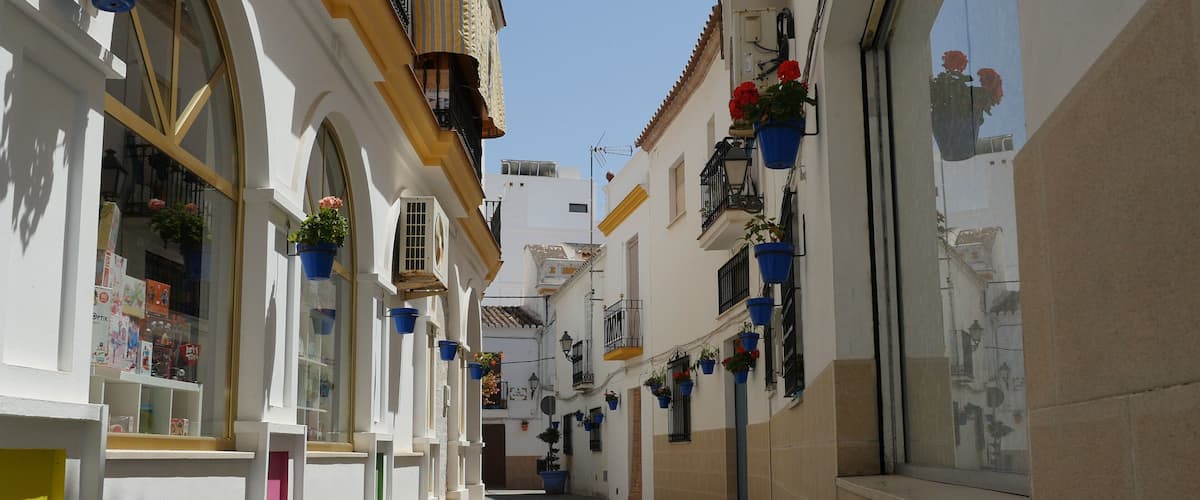 A narrow cobblestone street lined with white buildings, blue hanging flower pots, and red flowers.