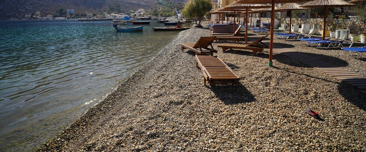 A pebbled beach with wooden lounge chairs and thatched umbrellas, boats docked in the background, and mountains in the distance.