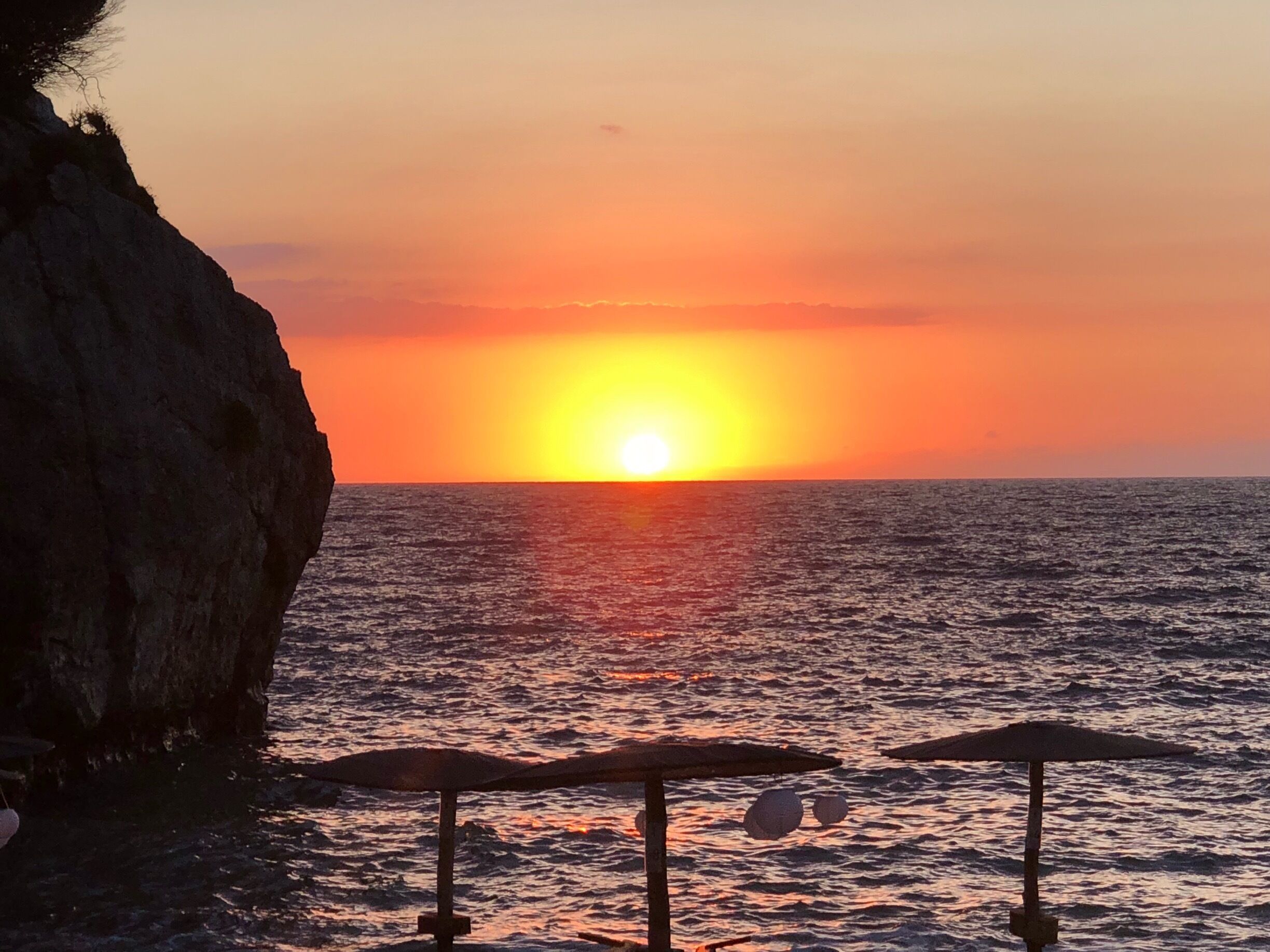A sunset over the ocean with a rocky cliff and beach umbrellas.